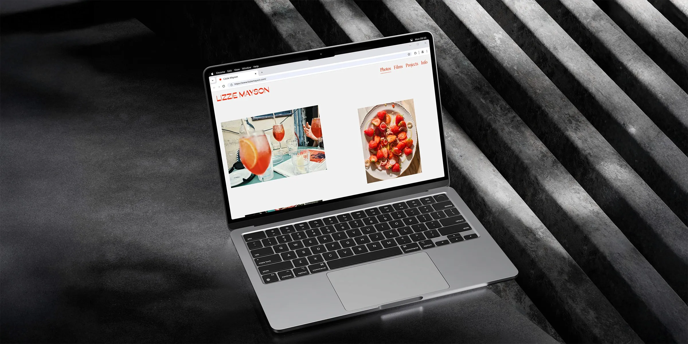 A sleek laptop displays a minimalist photography portfolio of Lizzie Mayson’s website, featuring vibrant images of cocktails and a close-up of strawberries, set against a modern, dark industrial background, highlighting a stylish creative workspace.